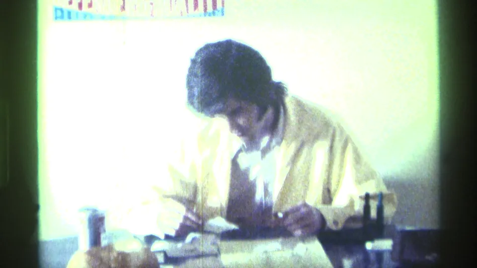 1976 Kentucky home-movie frame of an adult man seated at a table, looking down as he writes or handles papers.