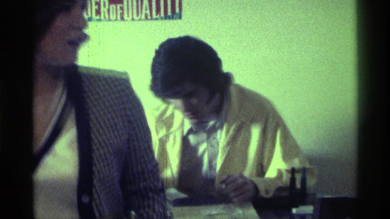 Man in a yellow jacket writes at a table while a woman stands partly in frame.