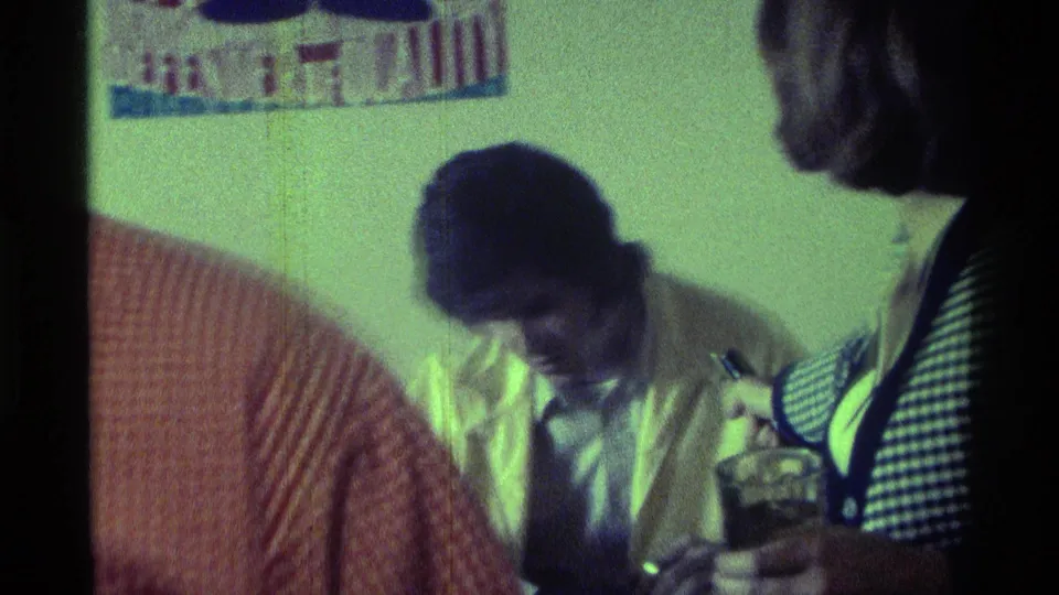 The seated man bends toward the tabletop while the woman at his side gestures with a small object near glasses and a can.
