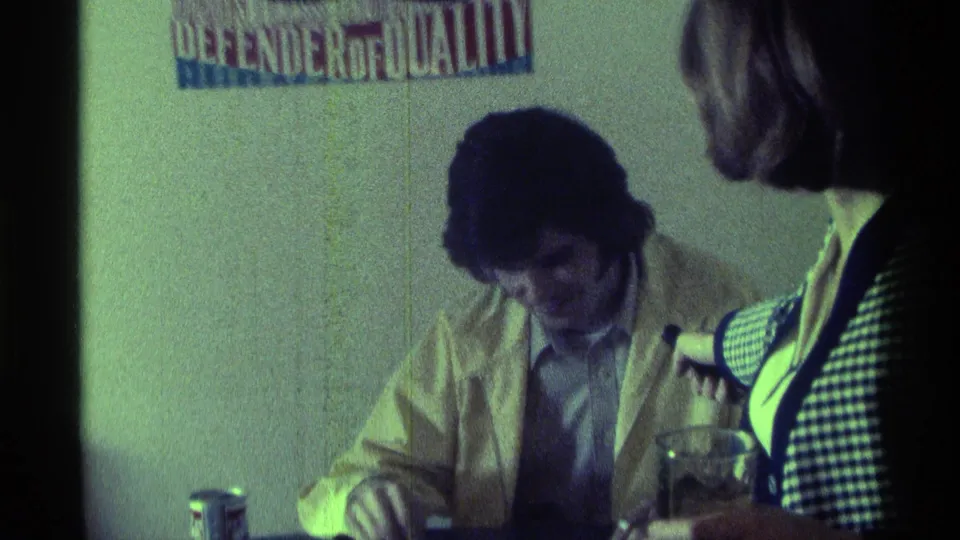 A man in a yellow jacket sits under a wall poster while a woman stands beside the table with glasses and a can visible in the foreground.