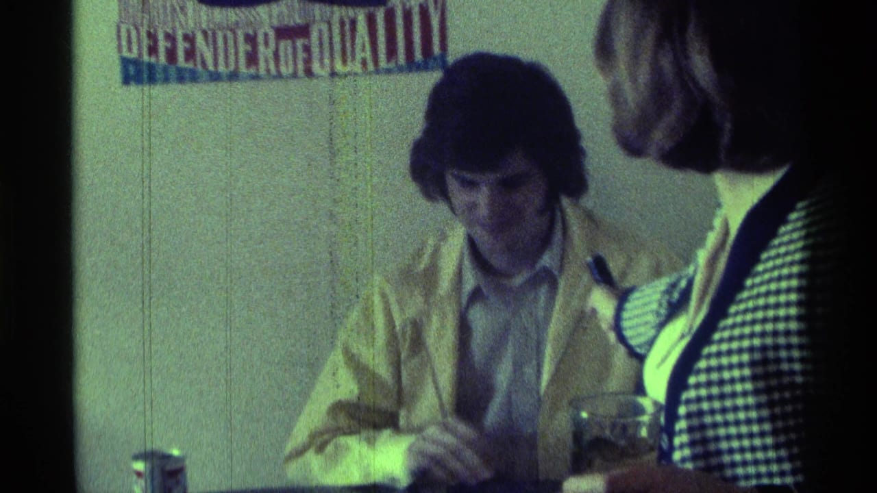In 1976 Kentucky home-movie footage, a seated man in a yellow jacket faces a black microphone near a wall poster.
