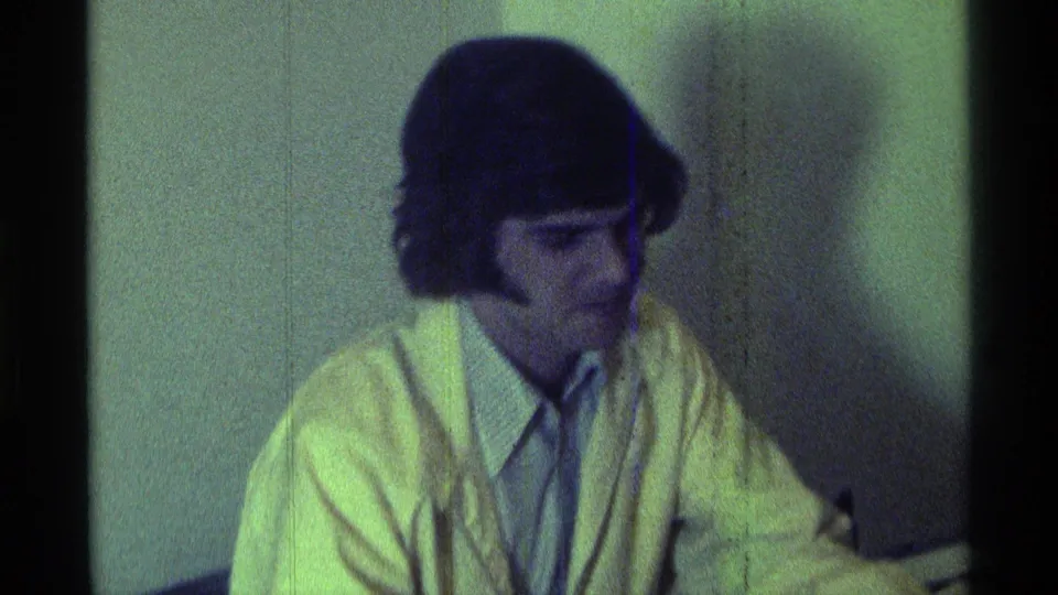A closer 1976 Kentucky 8mm view shows the seated man in a yellow jacket with a microphone held just below his face.