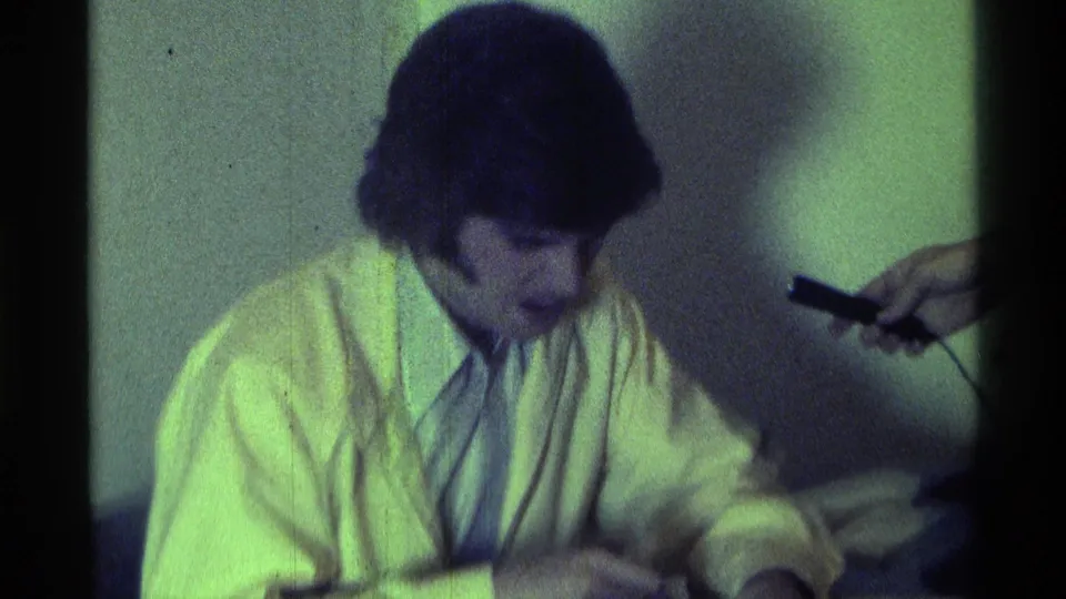 The man in a yellow jacket leans toward the table in this 1976 Kentucky interior as a black microphone enters from the right.