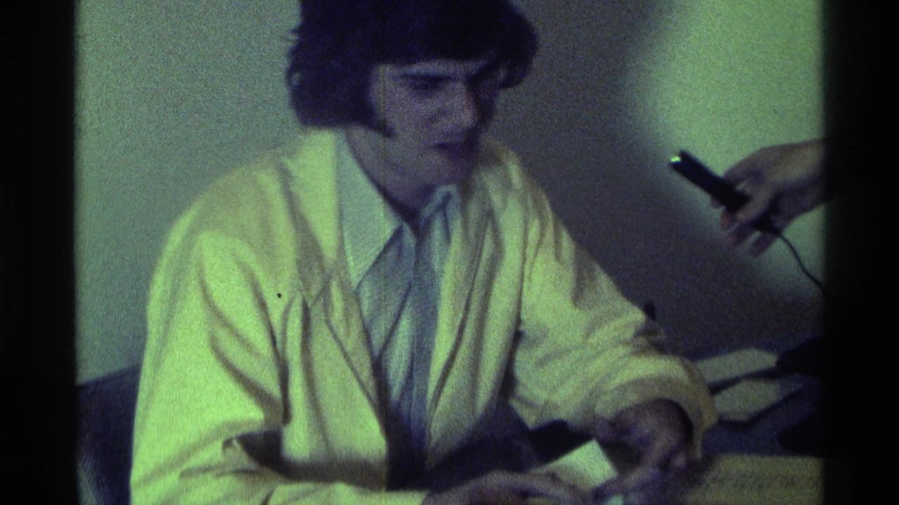 Seated man in a light jacket faces a handheld microphone at a table.