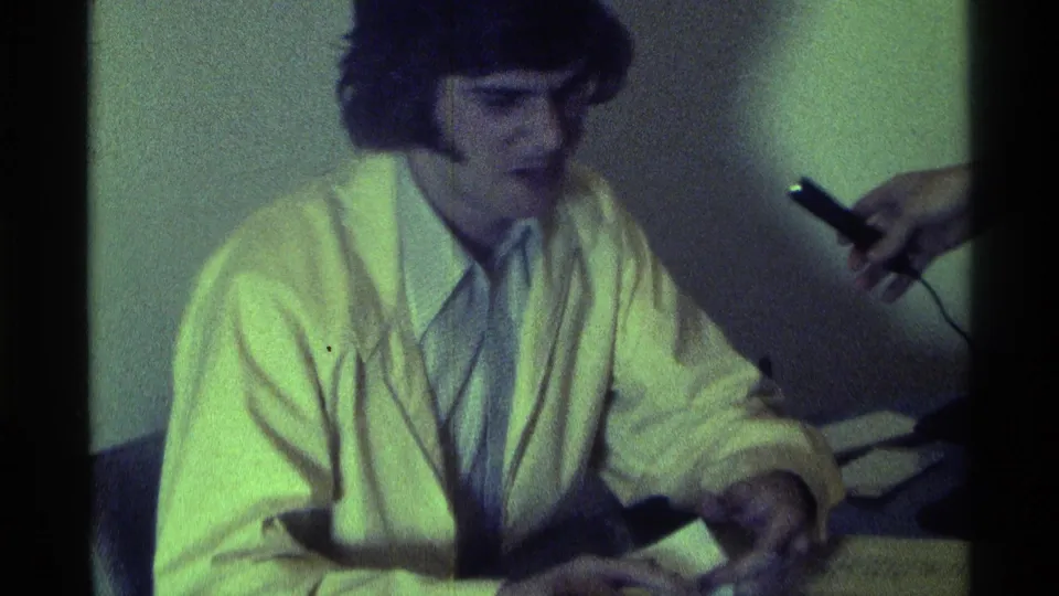 1976 Kentucky 8mm frame of a seated man with sideburns in a light jacket as an off-screen hand holds a microphone near him.