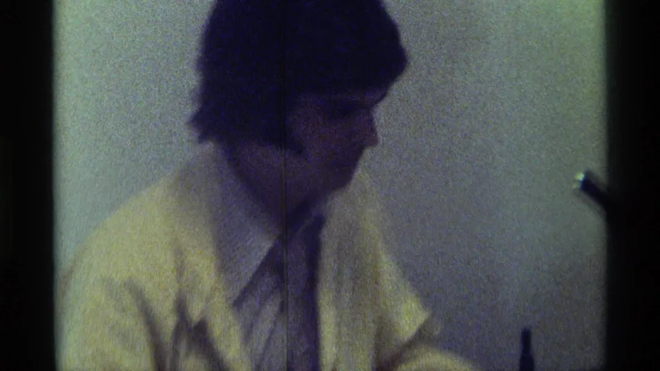Soft 8mm side view from 1976 Kentucky of the seated man in a light jacket during the indoor interview moment.
