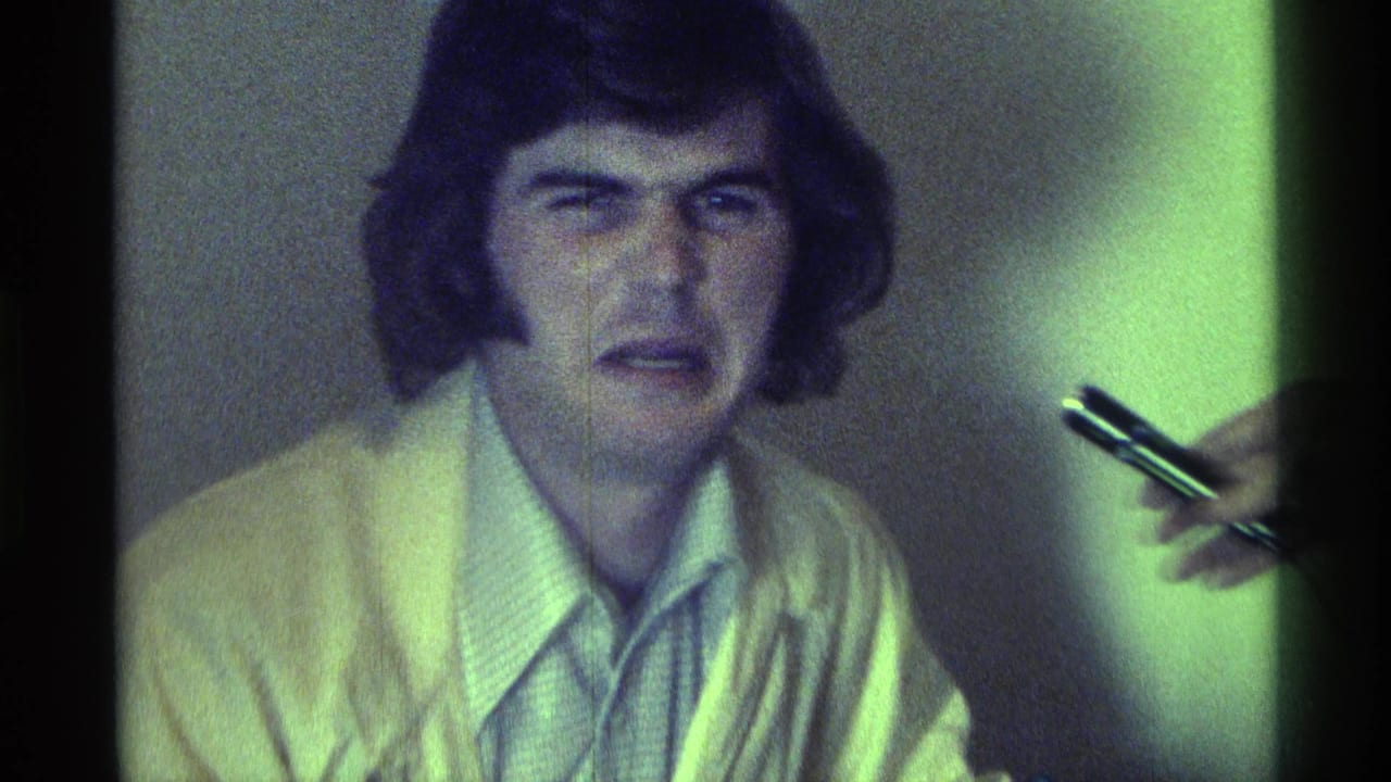 In 1976 Kentucky home-movie footage, a young man in a light sweater faces the camera beside a hand holding a pen.