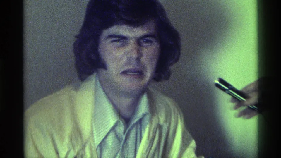 In 1976 Kentucky home-movie footage, a young man in a light sweater faces the camera beside a hand holding a pen.