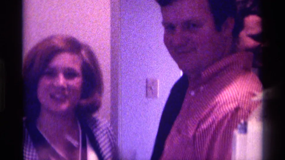 A woman smiles toward the camera as a man in a striped shirt turns beside her in 1976 Kentucky home-movie footage.