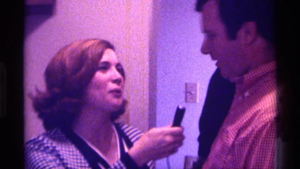 A woman in a checkered jacket appears to speak to a man in a striped shirt inside a 1976 Kentucky home interior.