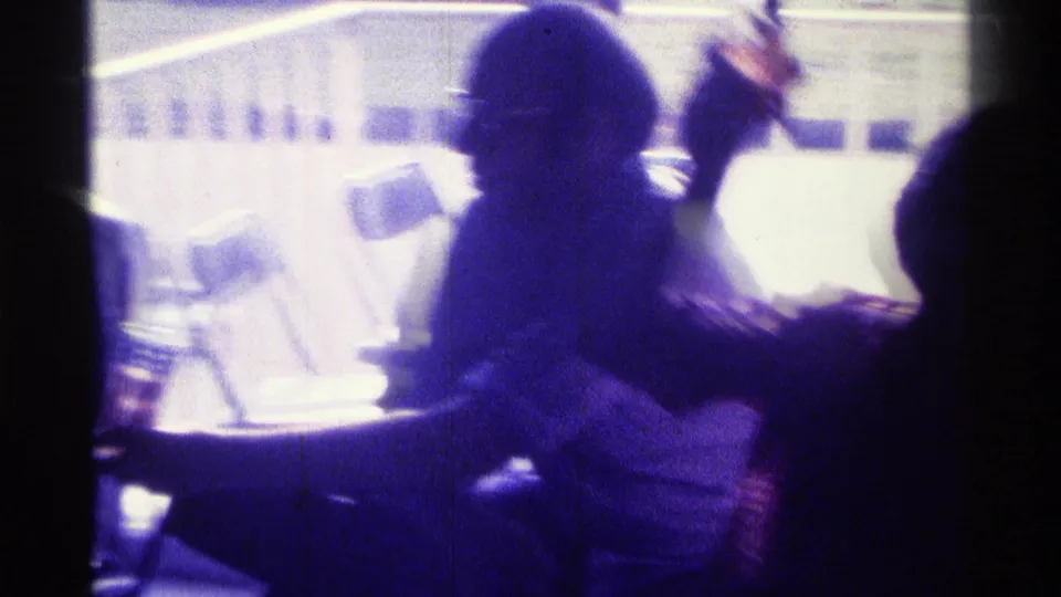 1976 Kentucky 8mm frame showing silhouetted adults in a dim private interior with strong blue-purple light and a table nearby.