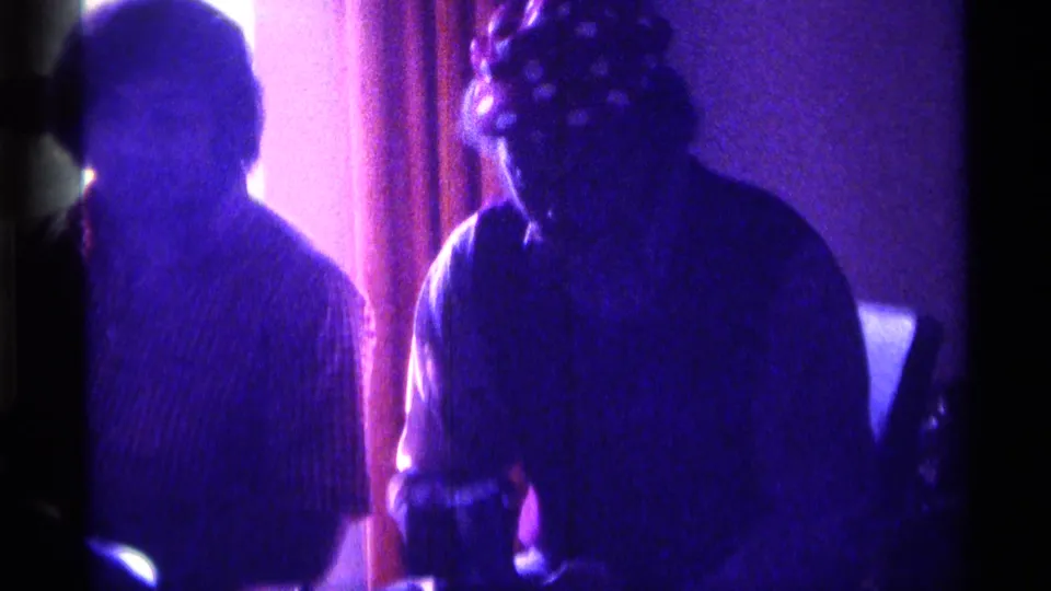 1976 Kentucky home-movie frame with two adults in a dark private room, one in a patterned shirt and one in darker clothing.