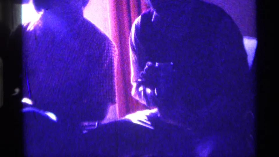 1976 Kentucky 8mm frame showing adults in low light, with one person holding a small object near a table in a private interior.