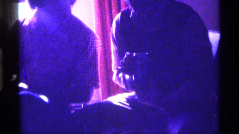 1976 Kentucky home-movie frame of adults gathered in a dim private room, with blue-purple exposure and a bright light behind them.