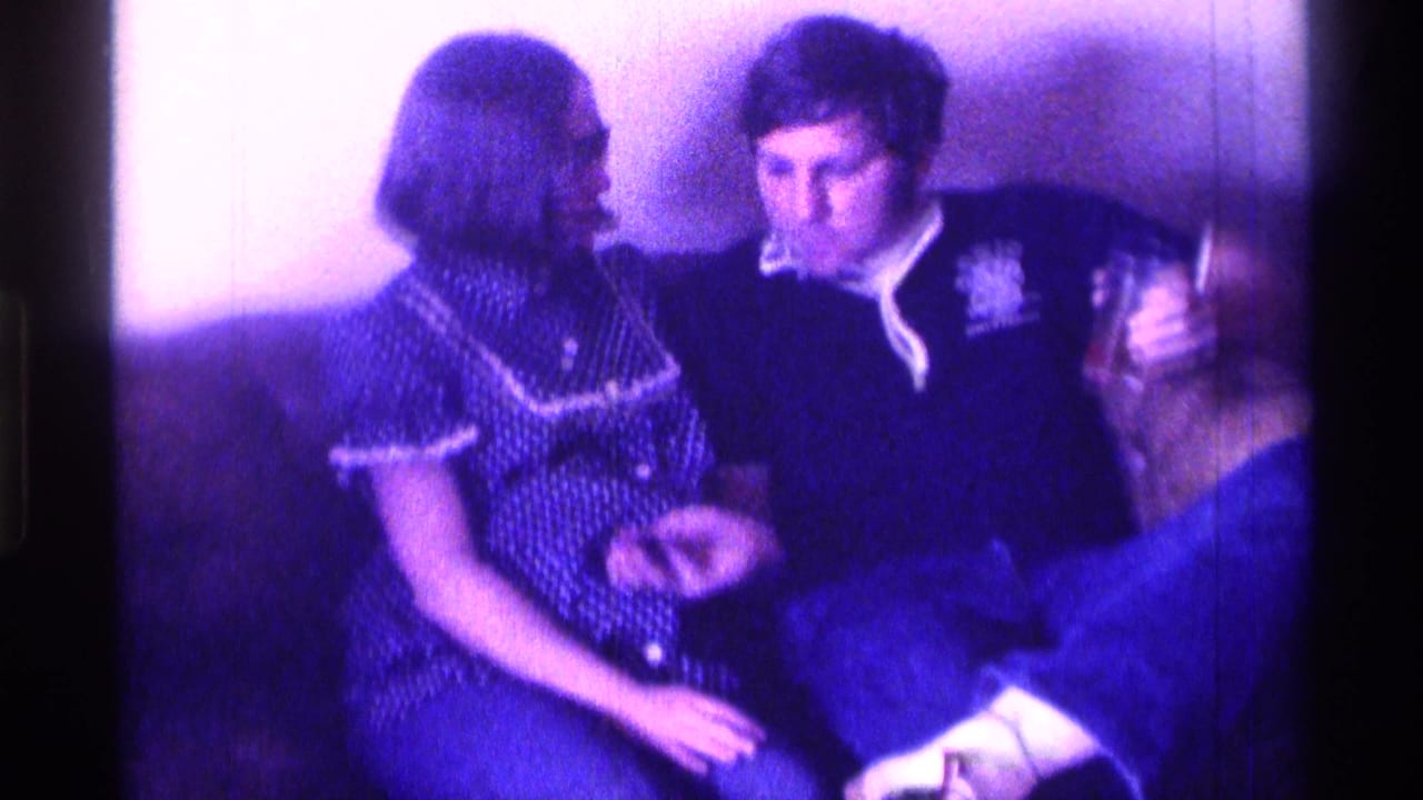 In a purple-tinted Kentucky 1976 home-movie frame, a young couple sits close on a couch while the man in a dark sweatshirt holds a glass.