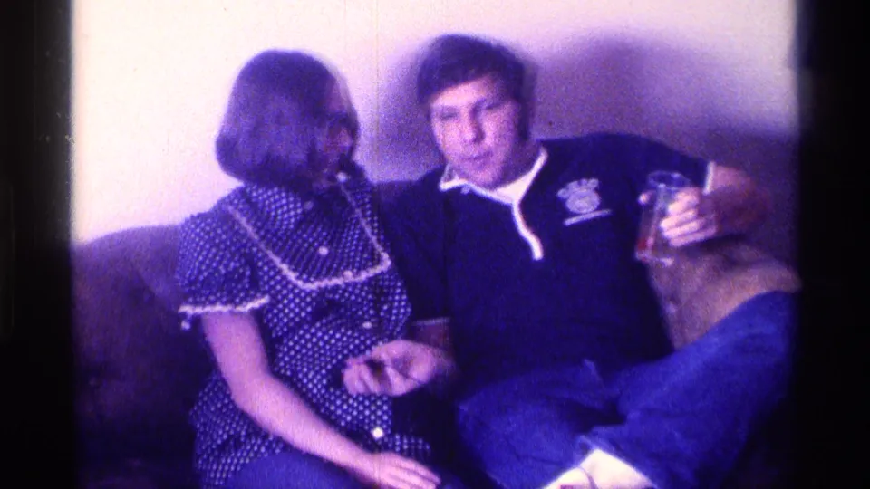 A young woman in a patterned dress and a young man in an Ohio University sweatshirt sit on a couch in a purple-tinted 1976 Kentucky home movie.