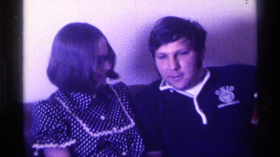 Close purple-blue home-movie frame of a young Kentucky couple on a couch in 1976, the woman turned toward the man in a university sweatshirt.