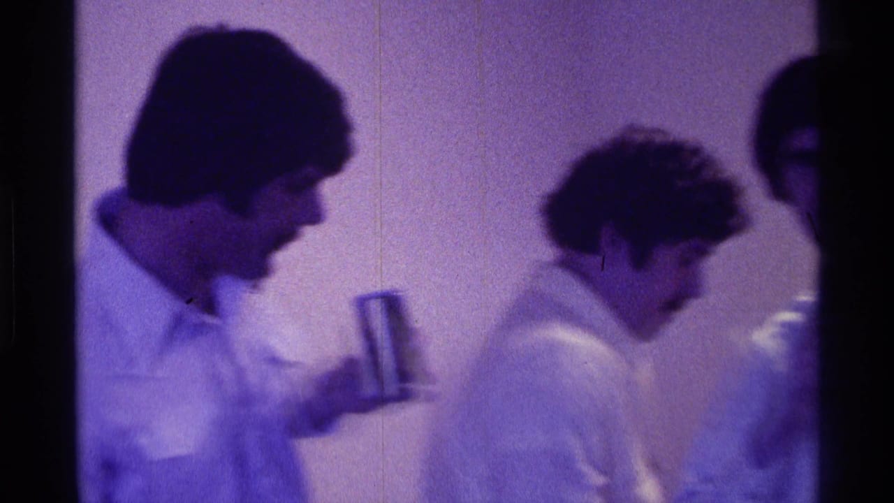 Young men in light shirts stand indoors as one holds a drink near a group.
