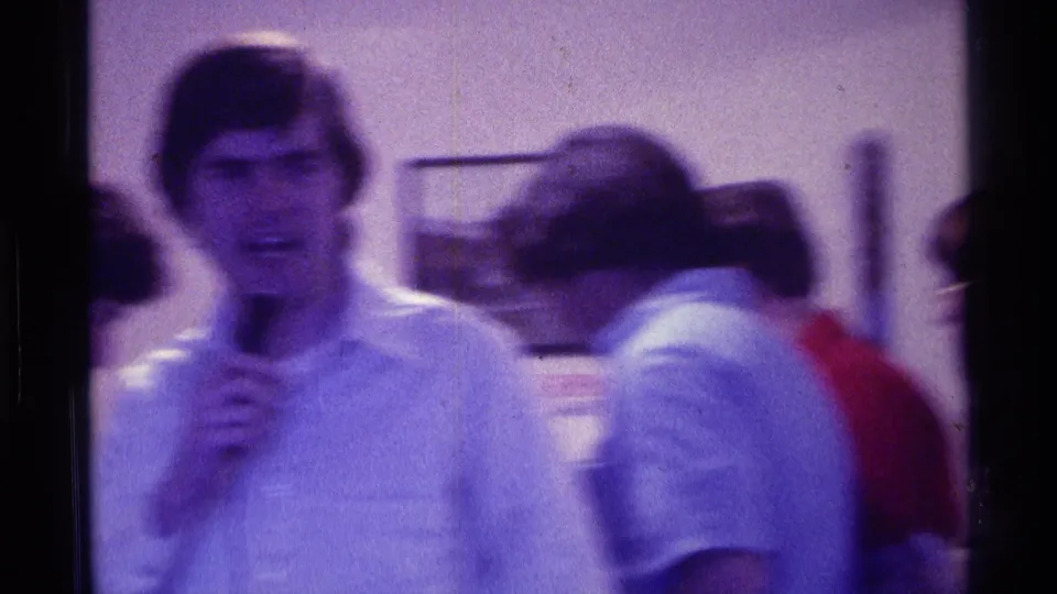 A young man in a light collared shirt holds a microphone at an indoor Kentucky gathering in 1976, with other attendees close by.