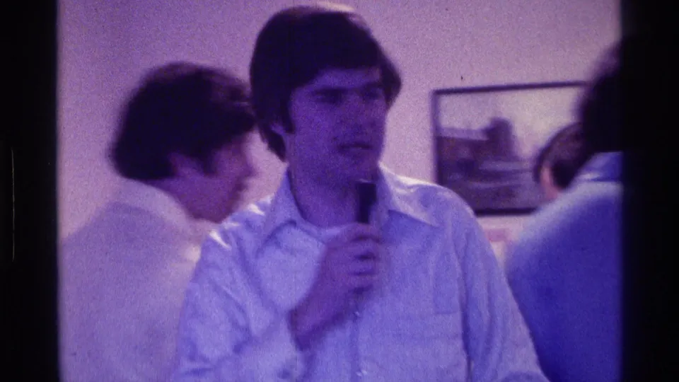 A bespectacled man holds a microphone in soft 8mm footage from a 1976 Kentucky indoor social gathering as people move behind him.