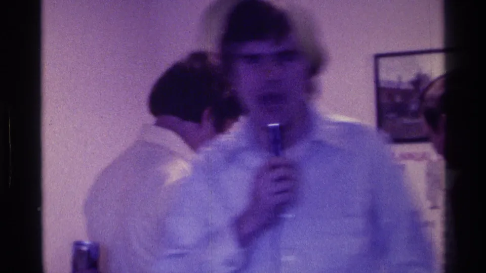 A soft-focused 1976 Kentucky home-movie frame shows a man with a microphone in front of nearby guests and wall art.