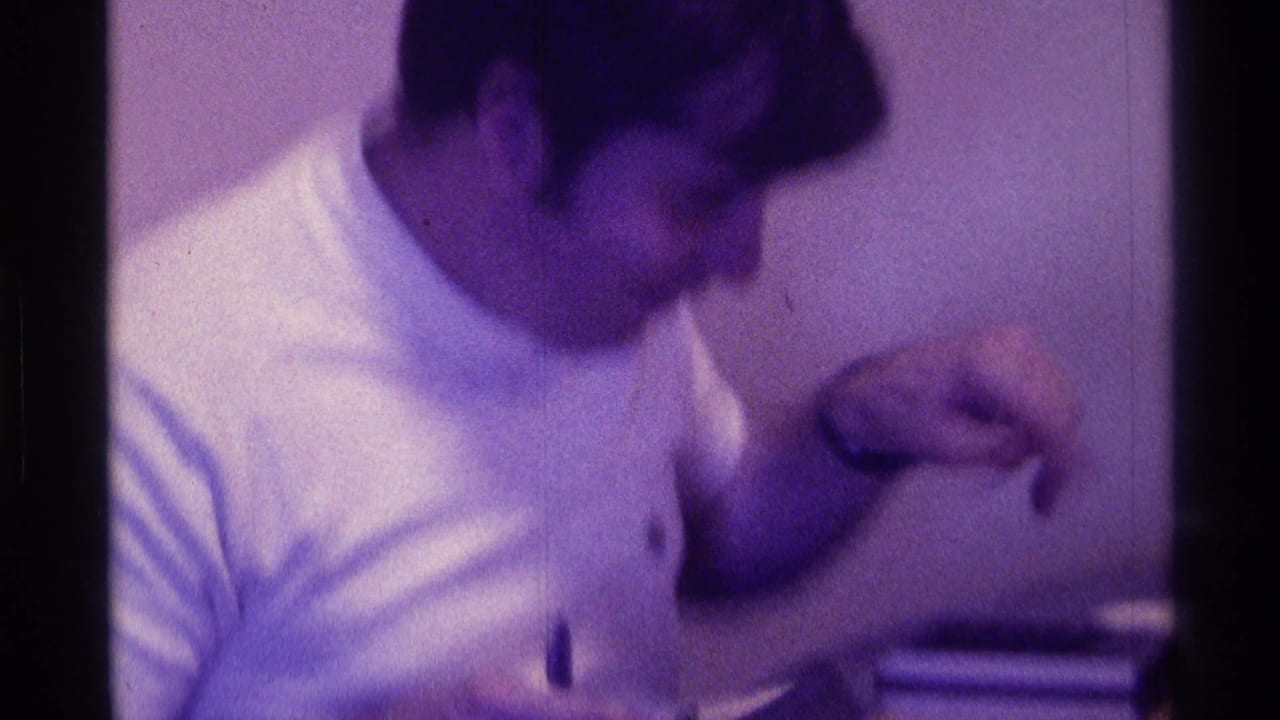 In this 1976 Kentucky home-movie frame, a young man in a white T-shirt sits indoors and looks down while gesturing with one hand.