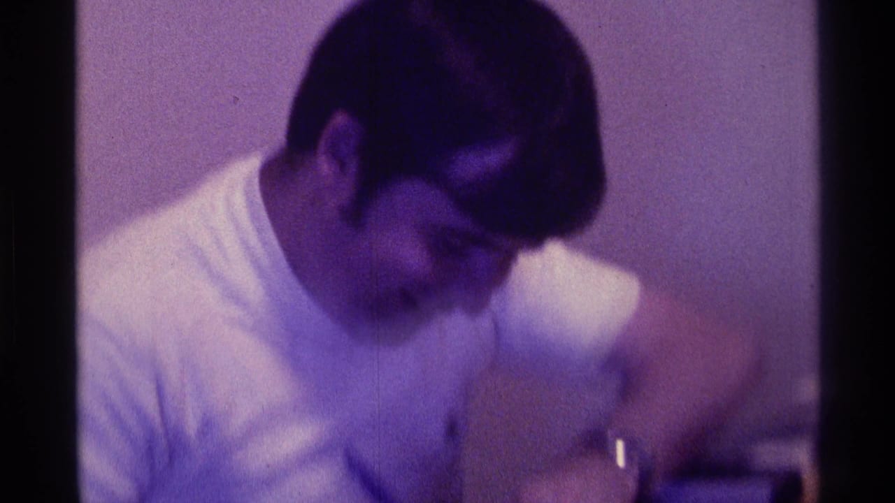 The man remains seated indoors in the Kentucky home-movie footage, leaning forward with his face turned down and one arm in motion.