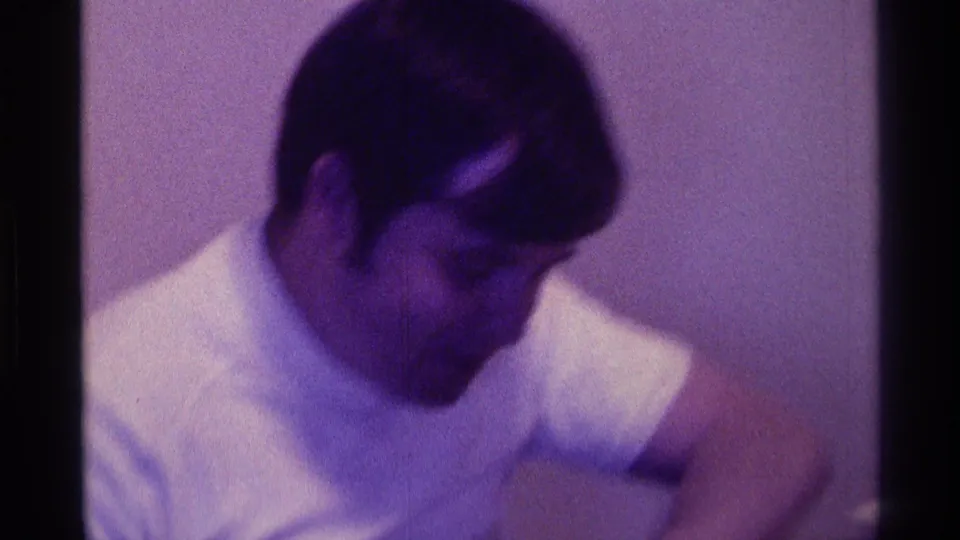 A soft 8mm frame shows the young man seated inside, head lowered, with one arm reaching or gesturing beyond the crop.