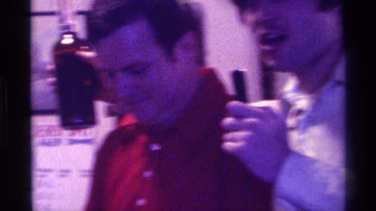 In 1976 Kentucky home-movie footage, two adult men stand indoors as the man in a red shirt holds a dark bottle.