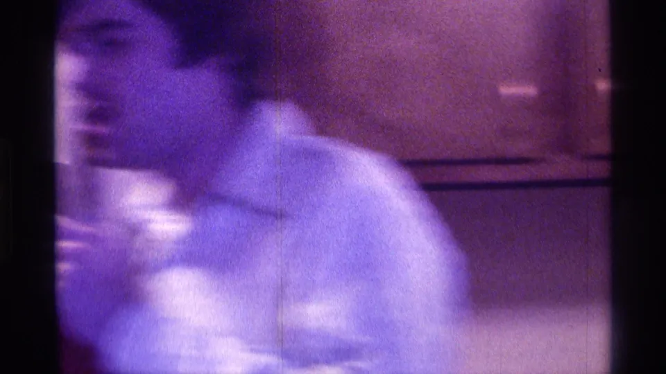 A man in a light collared shirt moves through a dim private interior in 1976 Kentucky home-movie footage.