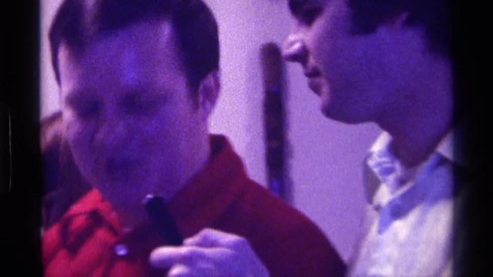 A man in a red shirt holds a dark bottle as another adult man stands nearby in a Kentucky indoor gathering.