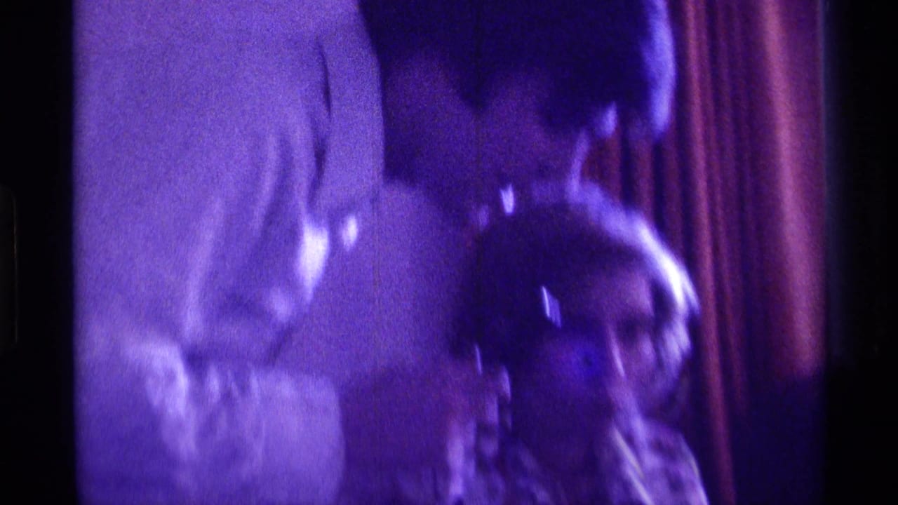 Blurred indoor frame with a man in a white shirt beside a seated woman in a floral blouse.