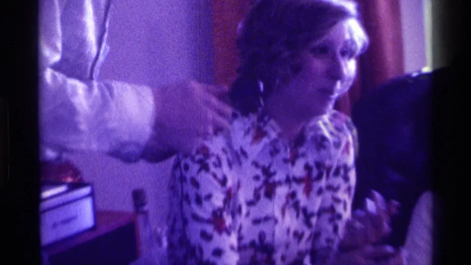 A woman in a floral blouse appears to answer while a man’s white-sleeved arm gestures beside her in 1976 Kentucky home-movie footage.
