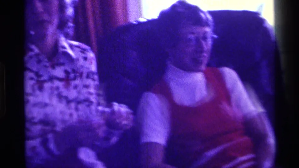 Two adult women sit in a private interior, with one wearing a floral blouse and another wearing a red vest over a white top.