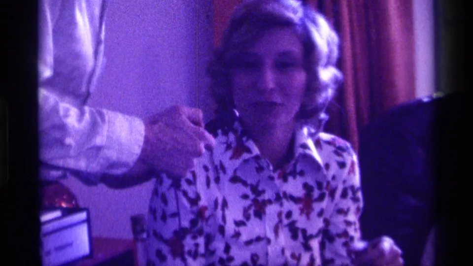 The woman in the floral blouse faces forward as a man’s arm reaches toward her with or near a microphone in purple-tinted 8mm footage.