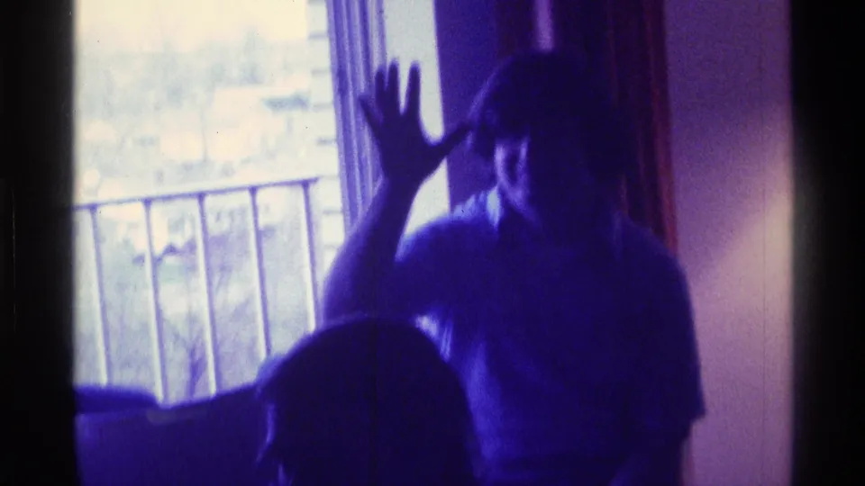 1976 Kentucky 8mm frame showing a man by a window raising his hand, with a seated figure partly visible in the foreground.