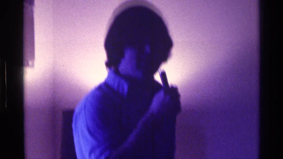 Purple-tinted 8mm frame from Kentucky showing a man in a collared shirt gesturing casually inside a private home.