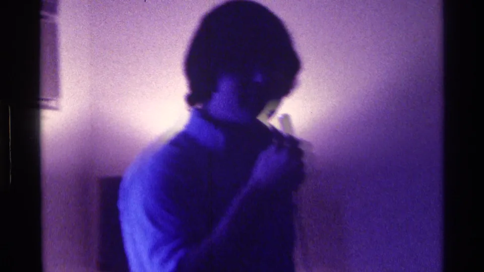 Young man at a 1976 Kentucky private gathering holds a microphone near his mouth under intense blue-purple light.