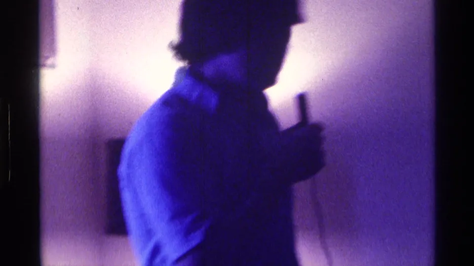 Side profile of a young man holding a wired microphone in a dim Kentucky home interior, filmed on 8mm in 1976.