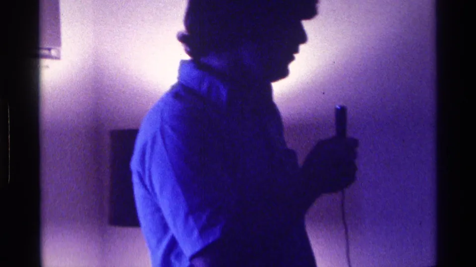 A 1976 Kentucky home-movie frame shows the young man in profile with a microphone against a glowing interior wall.