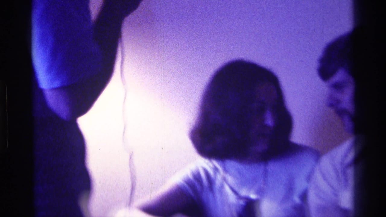 Young woman seated indoors with other people partly visible around her.