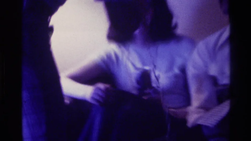 A young woman and a man sit close together indoors during a Kentucky 1976 home-movie social gathering.