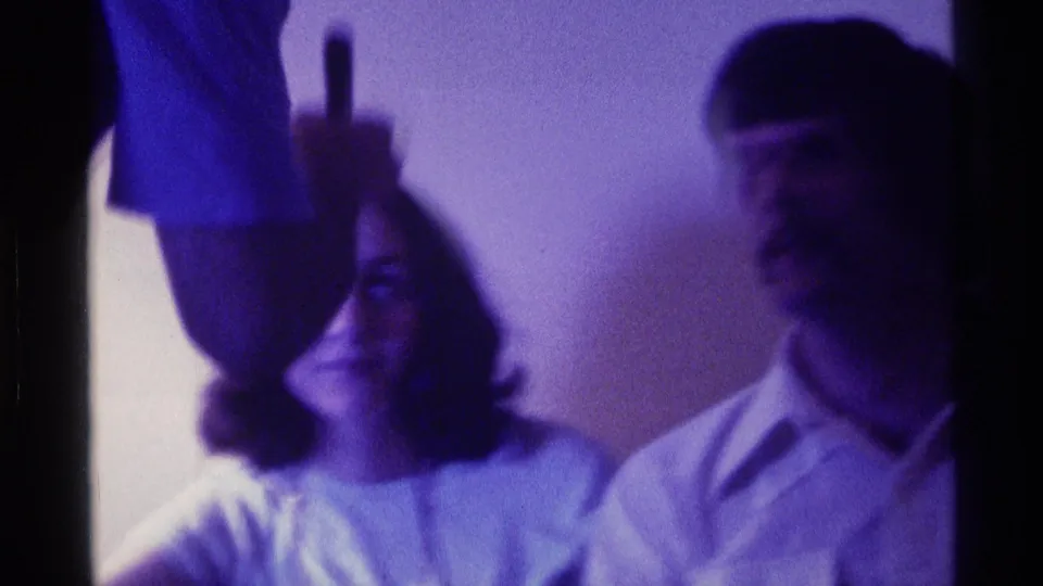 Two young adults sit indoors while a nearby hand holds a small dark object, captured with soft Super 8 focus and blue-purple color.