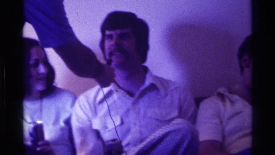 A young man sits with a drink as a nearby hand holds a microphone-like object near his face during the 1976 Kentucky gathering.