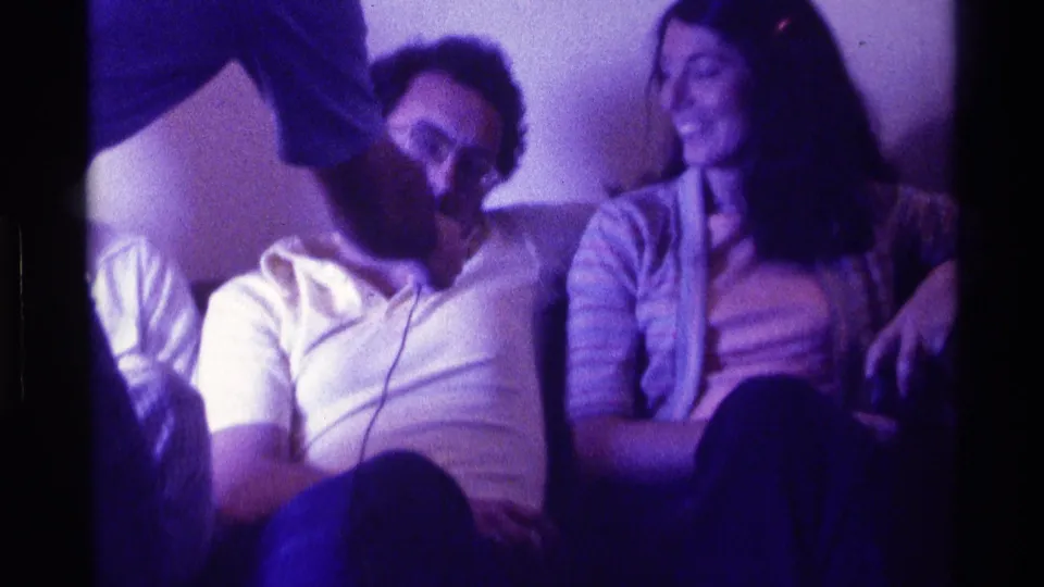 1976 Kentucky home-movie frame of young adults on a couch, with a microphone held toward a smiling group.