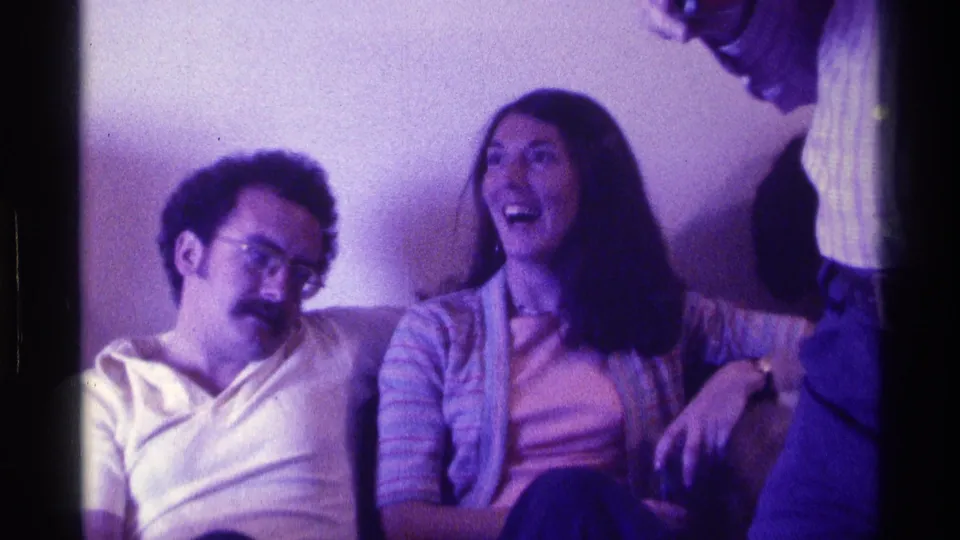 1976 Kentucky 8mm frame showing a man with glasses and a woman in a striped sweater seated on a couch during a casual indoor gathering.