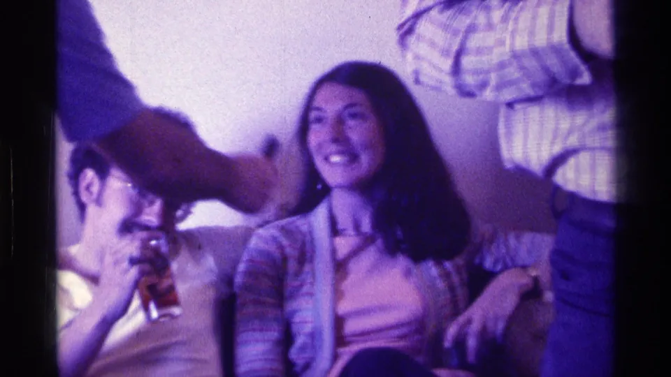 Purple-tinted 1976 Kentucky home-movie frame with a smiling woman on a couch and a man lifting a glass beside her.