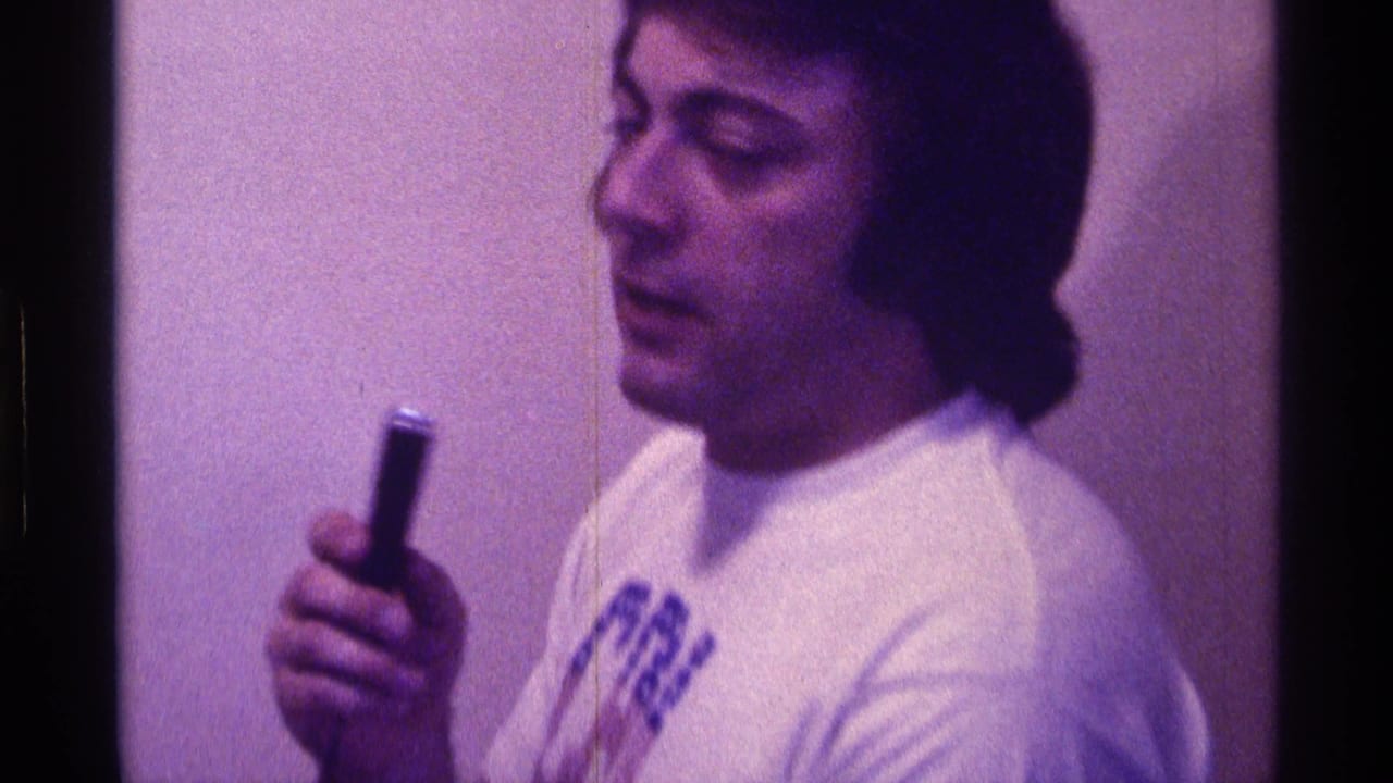 Man in a white graphic T-shirt holds a small microphone near his mouth in a dim interior.