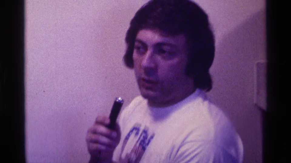 The man faces closer to the camera while holding a small microphone, with soft 8mm focus and purple-tinted indoor light.