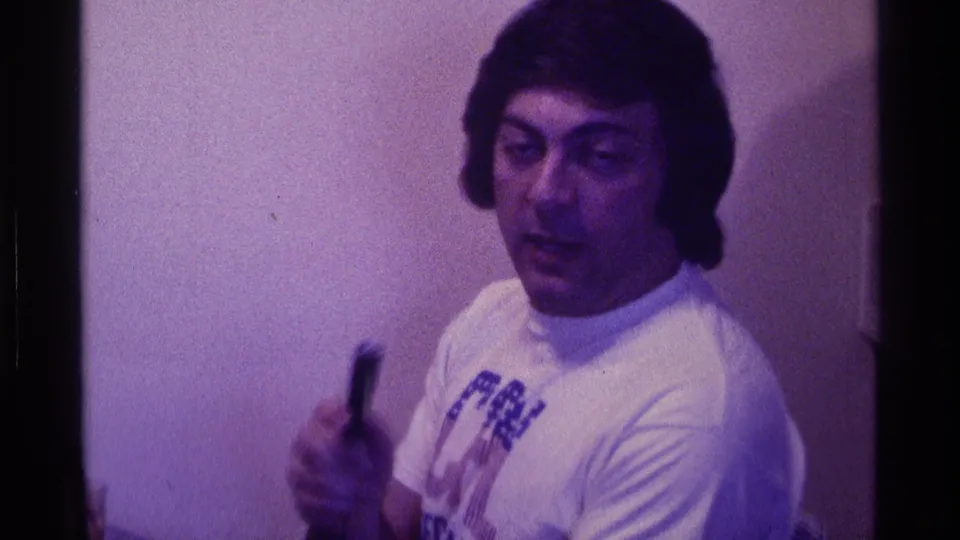 The man lowers the small microphone slightly and glances aside in the dim private interior, captured with visible 8mm grain.